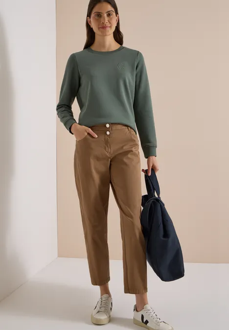 Femme en pull vert, pantalon beige et baskets blanches tient un sac bleu dans un espace lumineux.