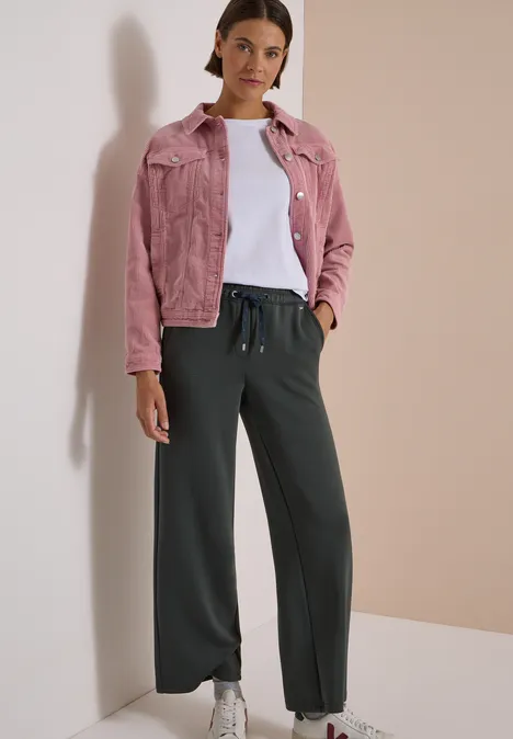 Spodnie z szerokimi nogawkami z tkaniny o fakturze przypominającej Silk Touch faded olive Kobieta na neutralnym tle w różowej kurtce jeansowej, białej koszulce, szerokich czarnych spodniach i białych trampkach.