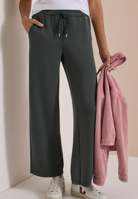 Spodnie z szerokimi nogawkami z tkaniny o fakturze przypominającej Silk Touch faded olive Kobieta ma na sobie szerokie, ciemnoszare spodnie dresowe z troczkami, białe trampki i trzyma różową bluzę.