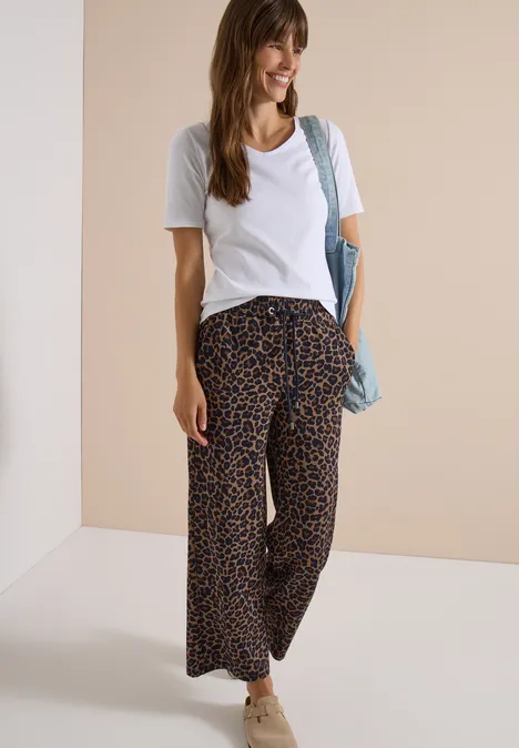 Broek met Wide Leg pijpen van Silk-Touch-kwaliteit creamy mocha Frau mit weißem T-Shirt, weitem Leopardenhose, Sneakern und Schultertasche, lächelnd.