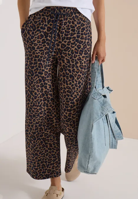 Broek met Wide Leg pijpen van Silk-Touch-kwaliteit creamy mocha Eine Person trägt weite Leopardenhose und helle Sneaker, hält eine Jeansjacke vor neutralem Hintergrund.