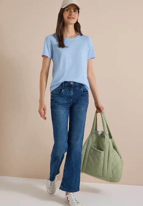 Frau in blauem T-Shirt und Jeans, weiße Sneakers und grüner Beutel vor neutralem Hintergrund.