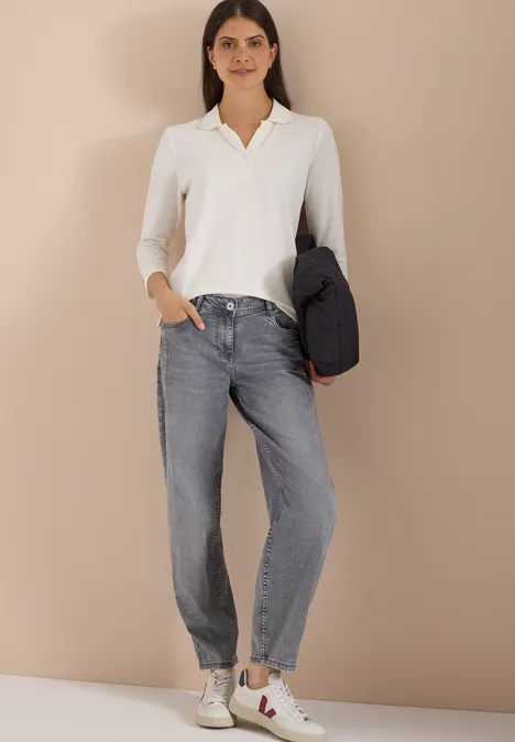 High Waist Barrel Leg Jeans w stylu Loose Fit light grey wash Młoda kobieta w białej koszuli, szarych dżinsach i trampkach, trzyma kurtkę pod ramieniem.