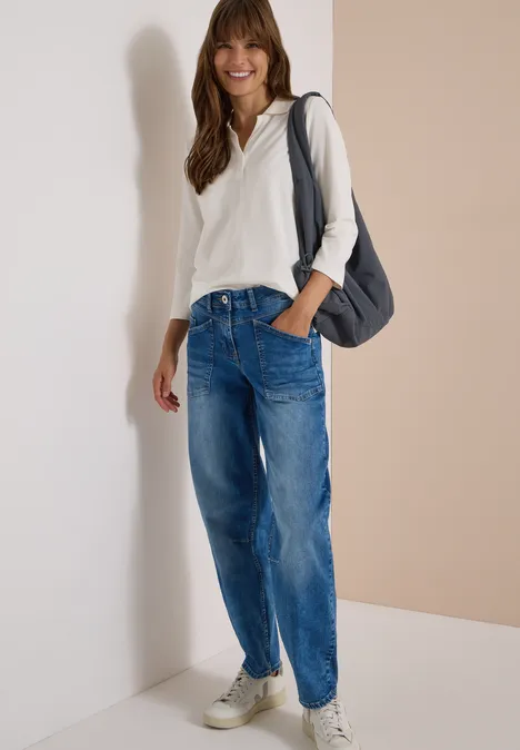 Jeans met High Waist en wijd uitlopende pijpen in een Loose Fit-pasvorm authentic used wash Vrouw in wit hoodie, losse jeans en sneakers, glimlachend tegen muur leunend met schoudertas.