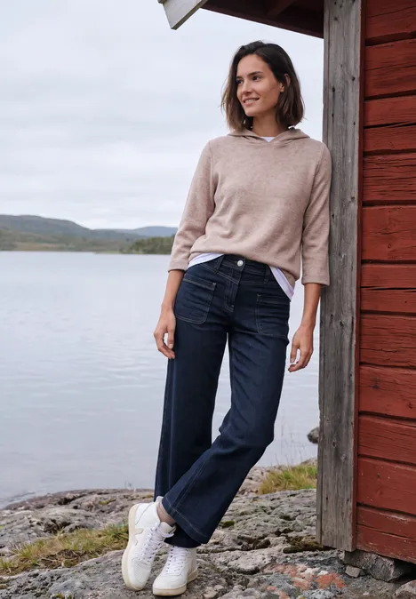 Wide Leg Jeans mit Patch-Pockets rinsed wash Frau lehnt lächelnd an roter Holzhütte am See, trägt Pullover, Jeans und weiße Sneakers, im Hintergrund Natur.