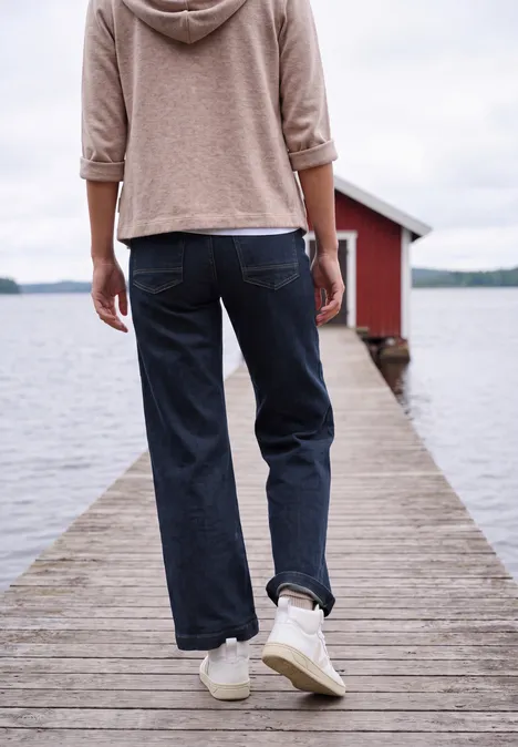 Wide Leg Jeans mit Patch-Pockets rinsed wash Person im Hoodie und Jeans läuft auf einem Holzsteg zum roten Bootshaus an einem ruhigen See.