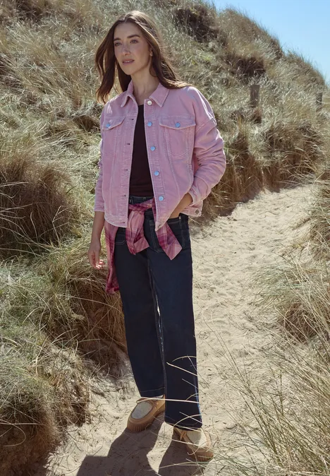 Wide Leg Jeans mit Patch-Pockets rinsed wash Frau in rosa Jeansjacke, weitem Dunkelblau-Jeans und beigen Sandalen läuft über einen grasigen Dünenpfad.