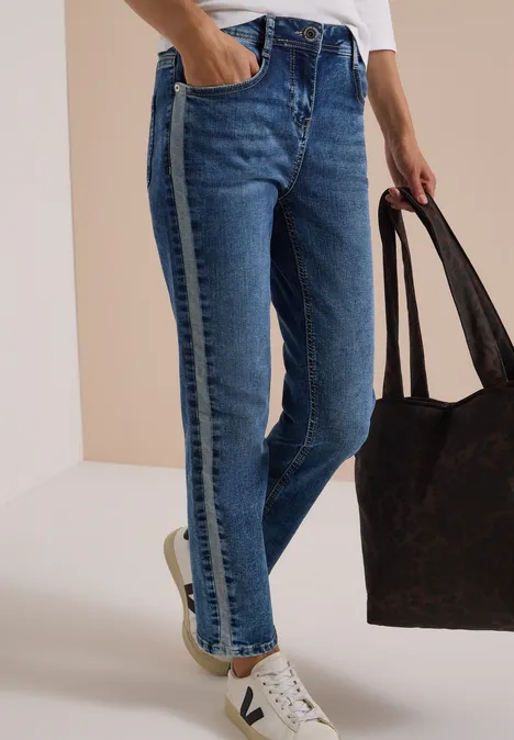 Eine Person trägt blaue Jeans mit seitlichem Streifen, weiße Sneaker und hält eine große braune Tasche.