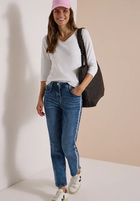 Lächelnde Frau in weißem Langarmshirt, blauer Jeans, weißen Sneakern und rosa Cap mit großer Tasche vor neutraler Wand.