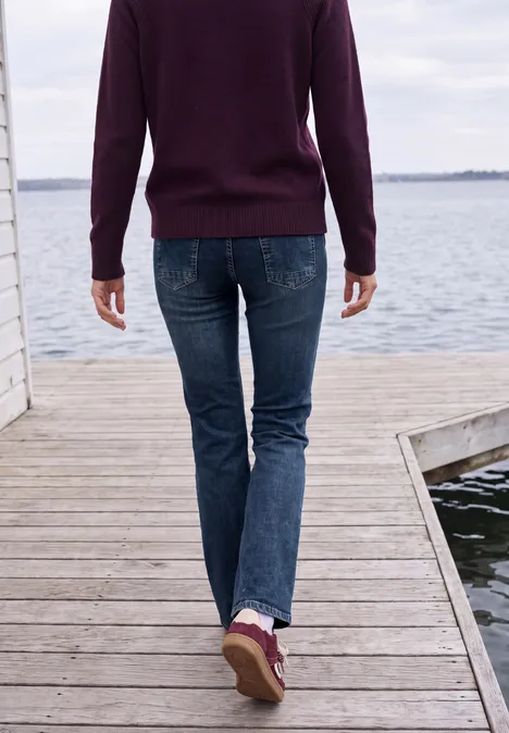 High Waist Bootcut Jeans im Slim Fit mid blue used wash Person in Jeans und Pullover läuft auf einem Holzsteg am Wasser, Blick von hinten.