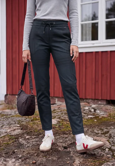 Mid Waist Slim Leg Hose im Casual Fit urban dark blue melange Person steht vor rotem Haus, schwarze Hose, weiße Sneaker mit rotem V, hält Tasche.