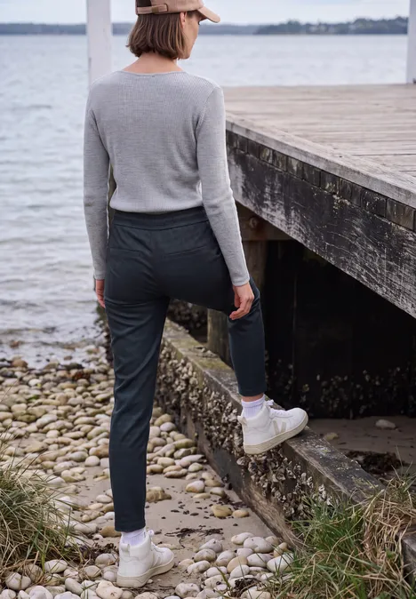 Mid Waist Slim Leg Hose im Casual Fit urban dark blue melange Person in grauem Oberteil, dunkler Hose und weißen Boots steht am Wasser neben einem Holzsteg.