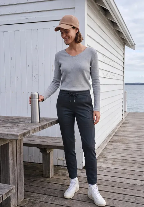 Mid Waist Slim Leg Hose im Casual Fit urban dark blue melange Frau in grauem Pullover, dunkler Hose und Cap steht auf einem Steg vor einem Schuppen am Wasser und hält einen Becher.