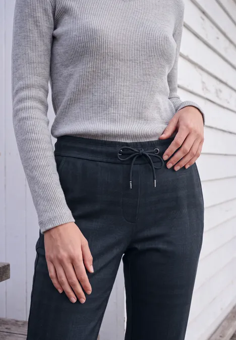 Mid Waist Slim Leg Hose im Casual Fit urban dark blue melange Frau in grauem Pulli und dunklen, taillierten Hosen mit Kordel vor weiß vertäfelter Wand.