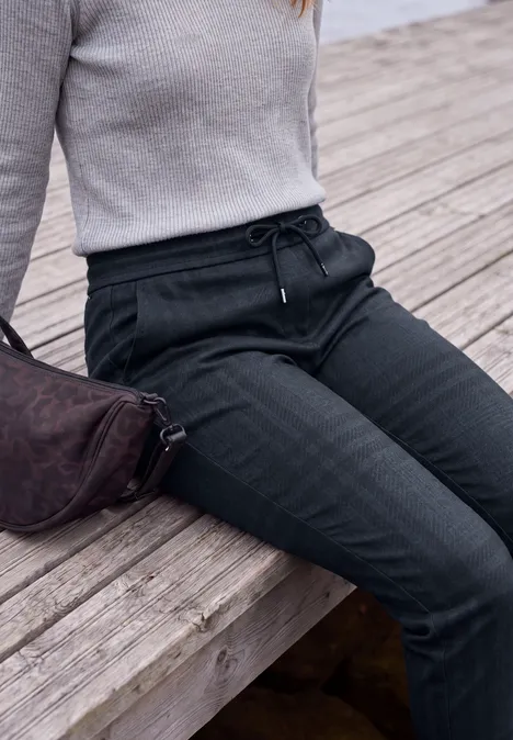 Mid Waist Slim Leg Hose im Casual Fit urban dark blue melange Person in grauem Pullover und schwarzen Jogginghosen sitzt auf hölzerner Bank am Wasser.