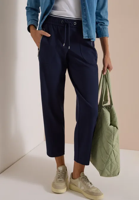 Frau trägt dunkelblaue, knöchellange Hose mit Tunnelzug, helle Sneaker und gesteppte grüne Tasche.