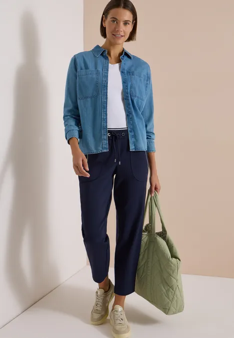 Frau trägt weißes T-Shirt, jeansblaues Hemd, dunkelblaue Hose, beige Sneaker und grüne, gesteppte Tasche.