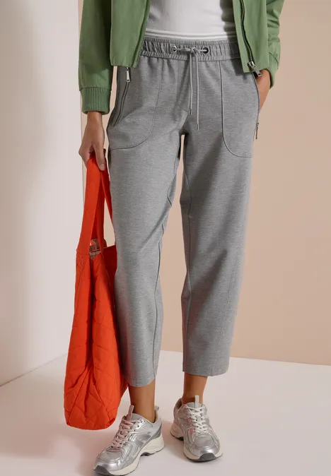 Frau trägt graue Jogginghose, silberne Sneaker, grüne Jacke, hält orangefarbene Tasche.