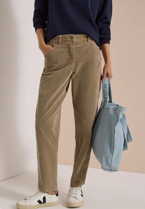 Mid Waist Barrel Leg Hose im Casual Fit creamy mocha Person trägt hellbraune Cordhose und weiße Sneaker, hält eine blaue Jeansjacke.