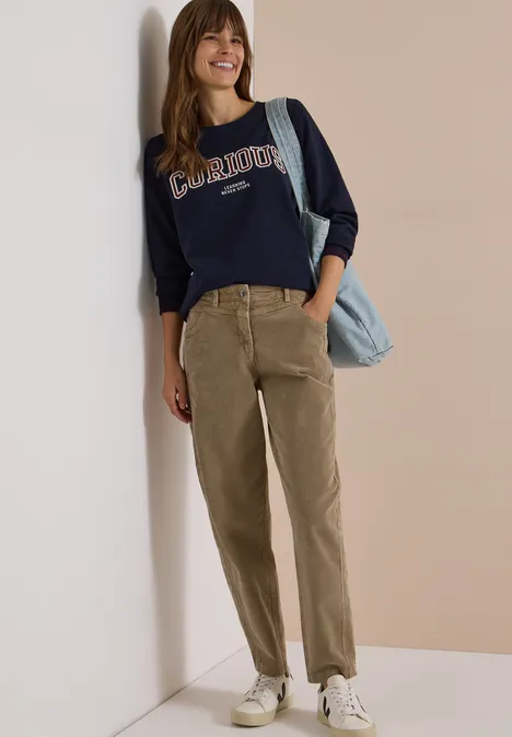 Mid Waist Barrel Leg Hose im Casual Fit creamy mocha Frau lehnt an Wand, lächelt, trägt dunkelblaues Sweatshirt, olivgrüne Cordhose, weiße Sneaker und blaue Tasche.