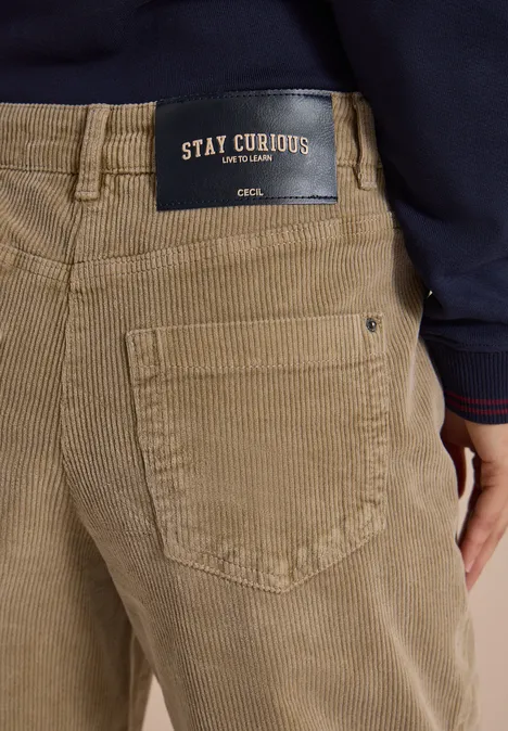 Mid Waist Barrel Leg Hose im Casual Fit creamy mocha Beige Cordhose mit Gesäßtasche und schwarzem Label 'STAY CURIOUS', darüber dunkelblaues Oberteil.