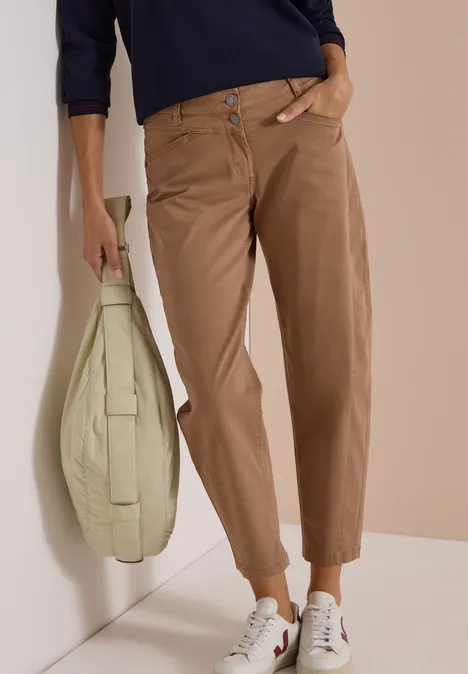 Een vrouw draagt een lichtbruine broek, witte sneakers, een donkerblauw shirt en een grote lichtgroene tas.