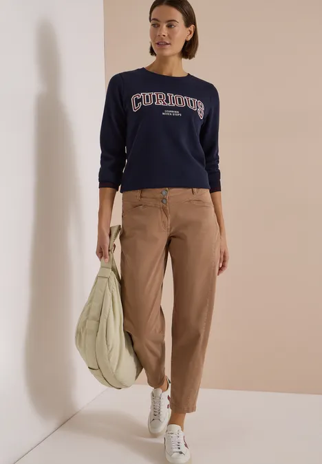 Vrouw in marineblauwe trui en beige broek staat voor lichte muur en draagt een gewatteerde tas.