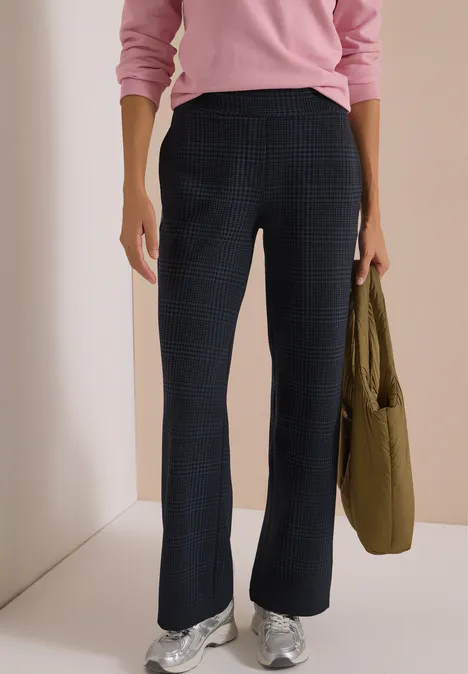 Vrouw met roze trui, zwarte broek en zilveren sneakers, met een groene tas in de hand.