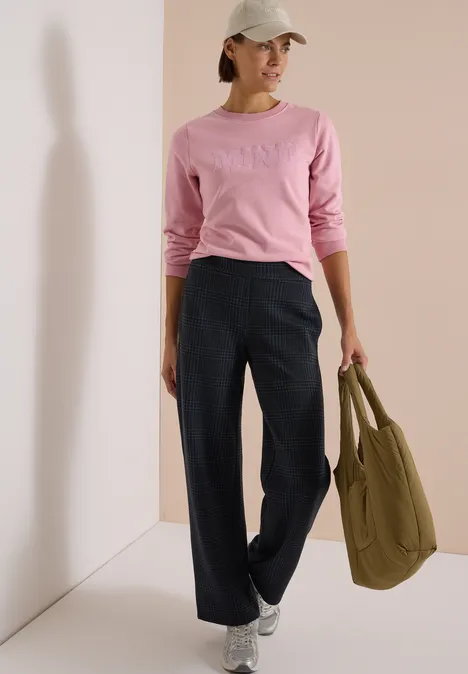 Vrouw in roze trui, wijde donkerblauwe broek, witte pet en sneakers, met kaki tas in hand.