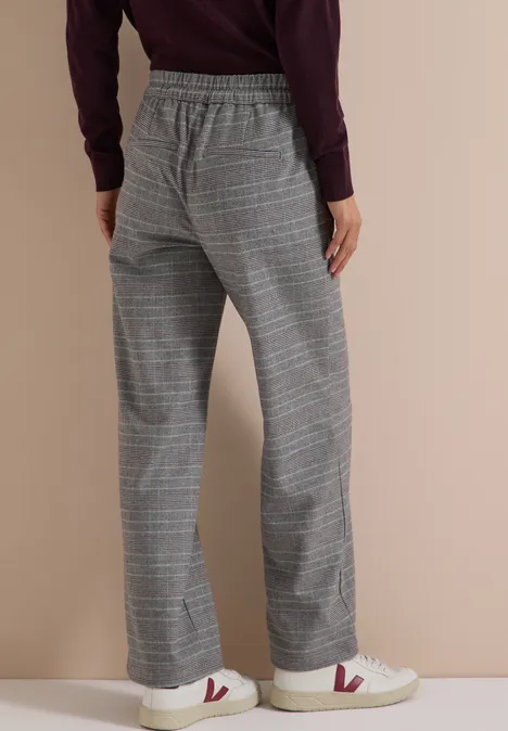 Vrouw draagt wijde grijze geruite broek en witte sneakers met rood V-logo.