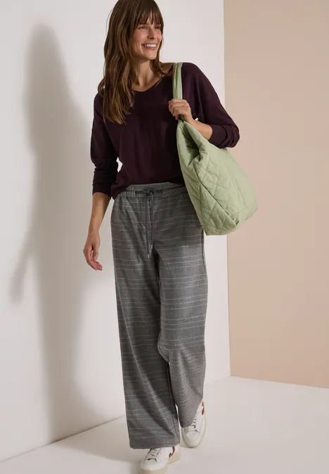 Vrouw in bordeaux sweater, grijze geruite wijde broek, witte sneakers en groen tas voor lichte achtergrond.