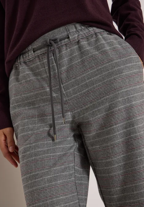 Close-up van een grijze geruite broek met trekkoord in de taille, gecombineerd met een bordeauxrode trui.