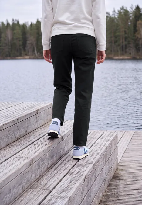 High Waist Barrel Leg Jeans im Loose Fit Black Person in schwarzer Hose und weißen Turnschuhen geht über eine Holzplattform am See.