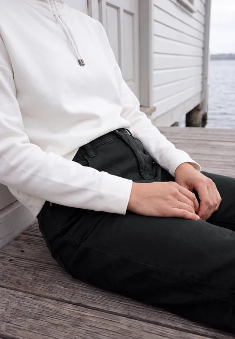High Waist Barrel Leg Jeans im Loose Fit Black Person sitzt auf Holzsteg am Wasser, trägt weißen Pullover und schwarze Hose.
