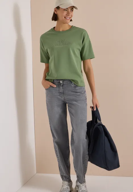 Frau in grünem T-Shirt, grauer Jeans und Cap, mit großer dunkelblauer Tasche vor beigem Hintergrund.