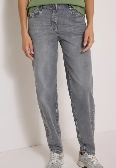 Person trägt gerade geschnittene graue Jeans und Sneaker, vor hellem Hintergrund stehend.