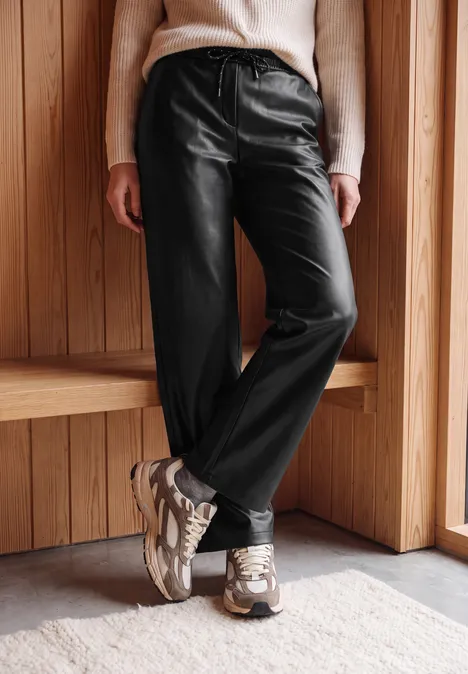 Broek met High Waist en wijde pijpen in leerlook Black Person trägt schwarze Lederhose, hellen Pullover und Sneaker, steht vor hölzerner Wand.