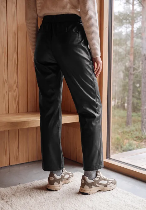Broek met High Waist en wijde pijpen in leerlook Black Eine Person trägt schwarze Lederhosen und Sneaker, steht vor einem Fenster mit Blick auf den Wald.