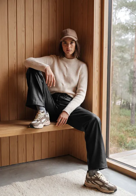 Broek met High Waist en wijde pijpen in leerlook Black Frau mit Cap, Pullover und weiten Hosen sitzt entspannt auf einer Holzbank am Fenster.