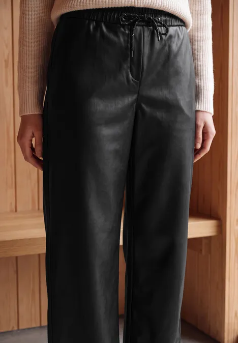 Broek met High Waist en wijde pijpen in leerlook Black Eine Person trägt schwarze Lederhose mit elastischem Bund vor einer Holzverkleidung.