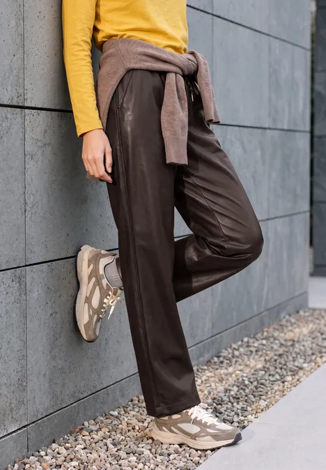 Broek met High Waist en Wide Leg pijpen in leerlook truffle brown Person lehnt an grauer Wand, trägt gelben Pullover, schwarze Hose, beige Sneaker und ein Sweatshirt um die Hüfte.