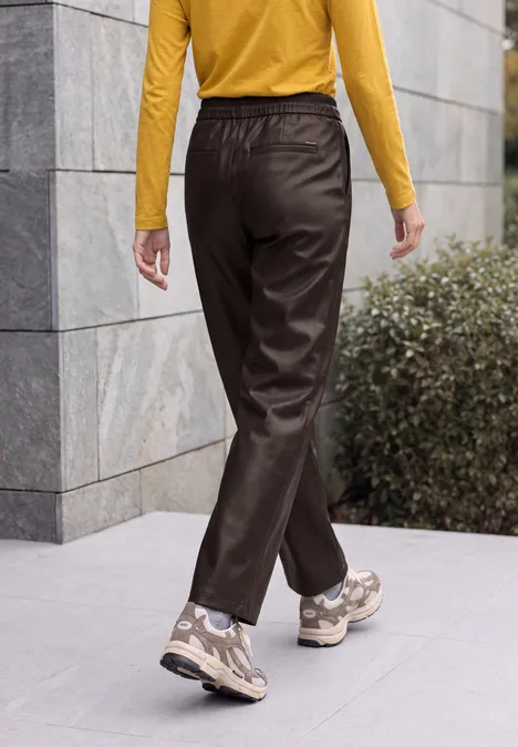 Broek met High Waist en Wide Leg pijpen in leerlook truffle brown Person in braunen Hosen, gelbem Oberteil und Sneakern, die an einer Steinwand auf einem Gehweg entlanggeht.
