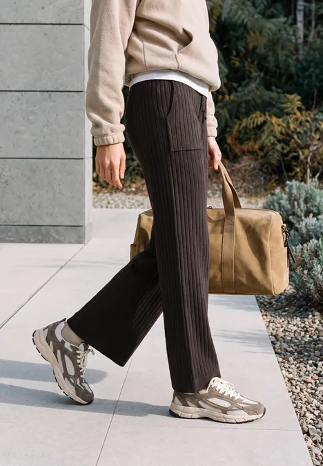 Person geht auf gepflastertem Weg, trägt braune Plisseehose, beige Sneaker und große Tasche.