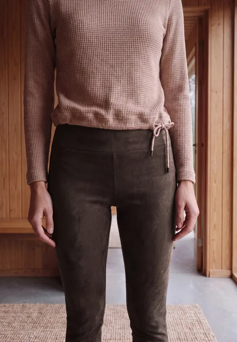 Une personne porte un pull marron et un pantalon marron foncé devant un mur en bois.