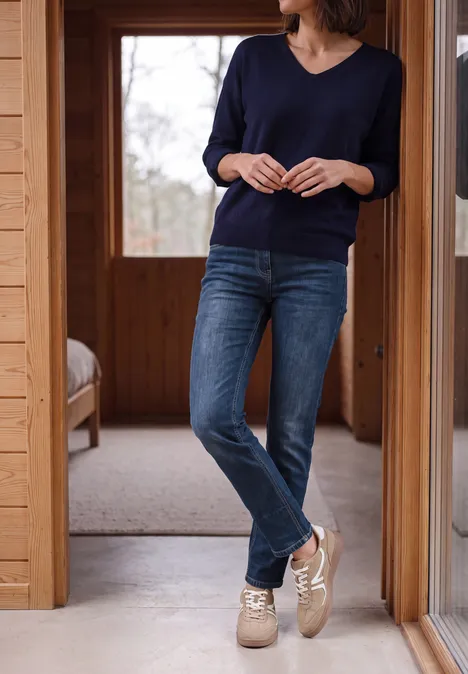 Eine Frau trägt Jeans, dunkelblauen Pullover und beige Sneaker in einem Holzhaus.