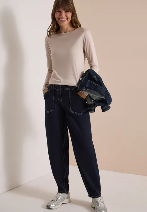 Vrouw in beige longsleeve, ruime spijkerbroek en sneakers, met een spijkerjack nonchalant over de schouder.