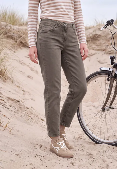 Person in olivgrünen Hosen, gestreiftem Langarmshirt und Sneakern steht im Sand neben einem Fahrrad.