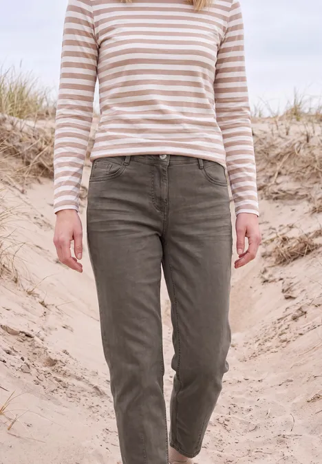 Person in beigeweiß gestreiftem Shirt und olivgrünen Hosen läuft über Sanddünen, Hintergrund mit Strandgras.