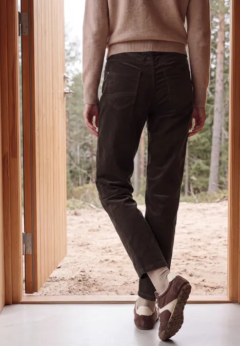 Eine Person steht in offen stehender Tür, trägt braune Hose, weiße Socken und robuste Schuhe, Blick in den Wald.