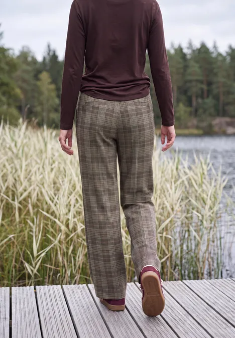 Person in karierter Hose und Langarmshirt steht auf einem Steg am See, umgeben von Schilf und Wald.